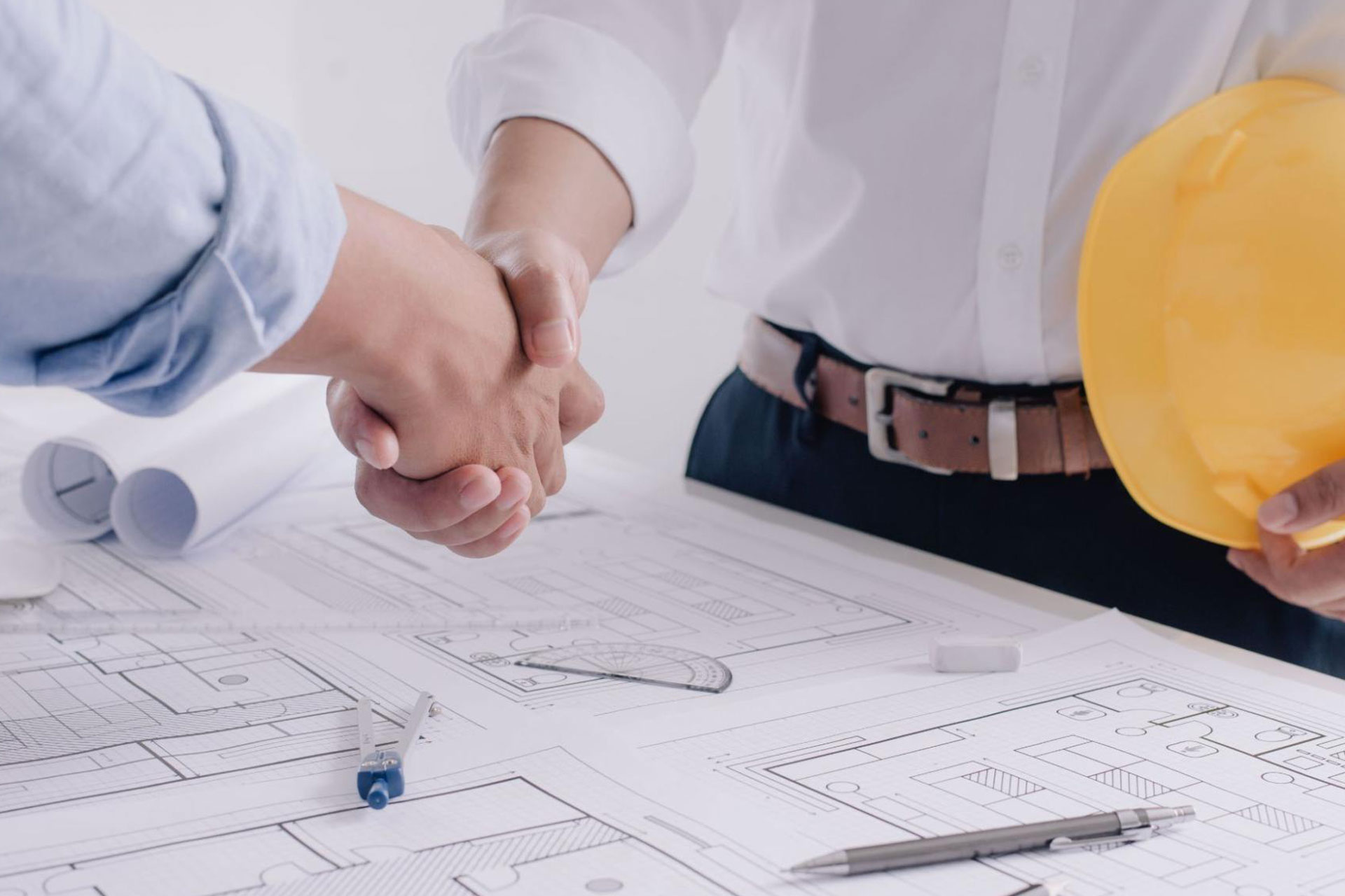 Two professionals shaking hands over a table covered with architectural blueprints, with one person holding a yellow hard hat. Two professionals shaking hands over a table covered with architectural blueprints, with one person holding a yellow hard hat.
