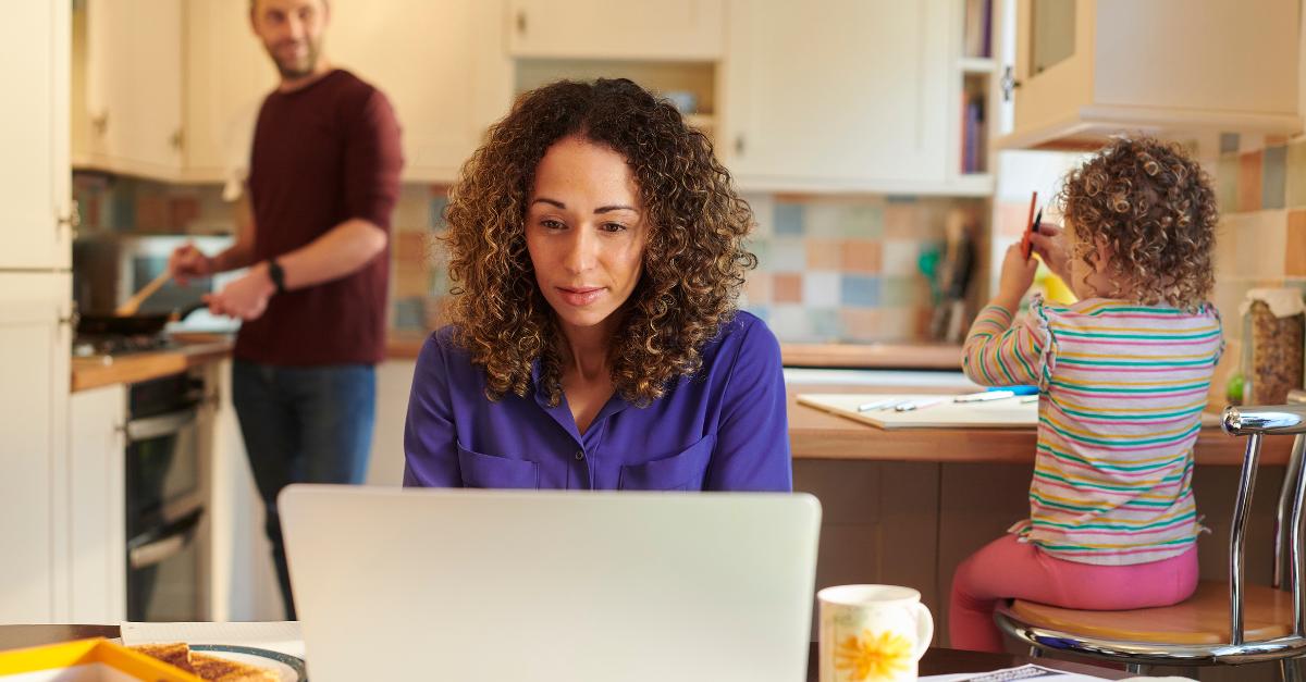 Woman working on laptop at home with partner and child behind her