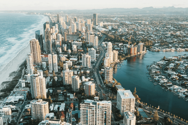Aerial view of the Gold Coast, Australia, featuring high-rise buildings along the coastline, a winding road, waterways, and residential areas stretching toward the mountains. Aerial view of the Gold Coast, Australia, featuring high-rise buildings along the coastline, a winding road, waterways, and residential areas stretching toward the mountains.